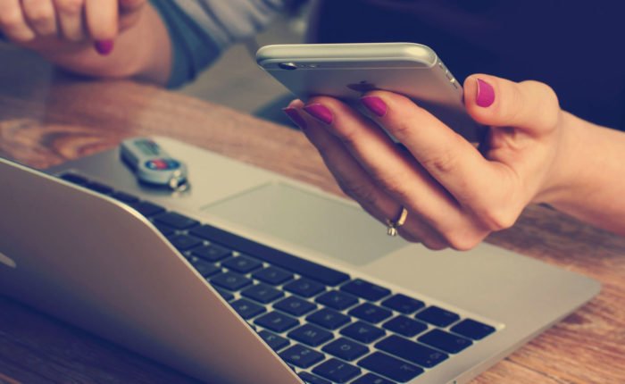 Woman with purple nails holds an Apple iPhone and sits at a laptop.