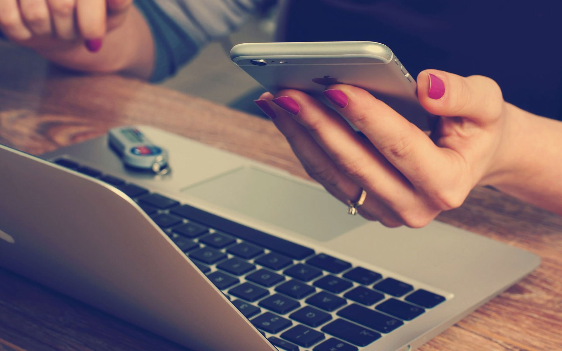 Woman with purple nails holds an Apple iPhone and sits at a laptop.