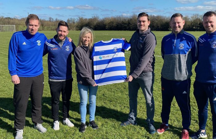 Bluescope's Ciarán Brogan and Deirdre Brogan hold a Wembley Rovers jersey with the Bluescope logo on it with the team management on a pitch.